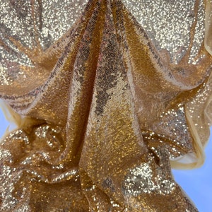 Puede incluir: Tela de lentejuelas doradas con un acabado brillante. La tela está drapeada y doblada, mostrando la textura de las lentejuelas.