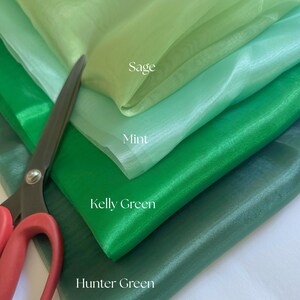 May include: A close-up of layered green fabric swatches in shades of sage, mint, kelly green, and hunter green. A pair of scissors with red handles is partially visible, suggesting the fabric is being cut or prepared for a project.