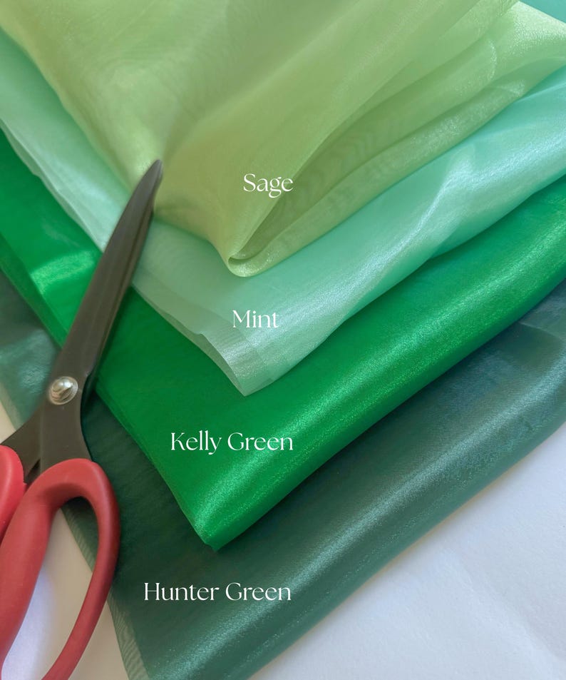 May include: A close-up of layered green fabric swatches in shades of sage, mint, kelly green, and hunter green. A pair of scissors with red handles is partially visible, suggesting the fabric is being cut or prepared for a project.