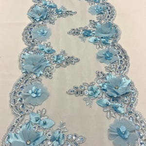May include: Light blue floral lace trim with 3D flowers, sequins, and pearls. The lace is on a sheer mesh background.