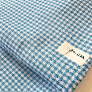 May include: Close-up of turquoise and white gingham fabric. The fabric is folded, revealing the pattern's repeating squares. A label with the word "TURQUOISE" is pinned to the fabric. The fabric is likely used for sewing, crafts, or home decor.