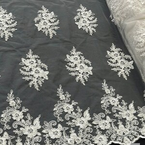 May include: White floral lace fabric with a scalloped edge. The lace features a repeating pattern of delicate flowers and leaves.
