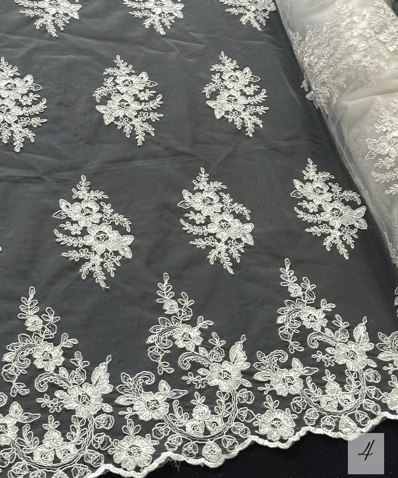 May include: White floral lace fabric with a scalloped edge. The lace features a repeating pattern of delicate flowers and leaves.