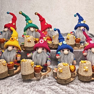 Puede incluir: Una colección de figuras de gnomos hechas a mano con largas barbas grises y coloridos gorros de ganchillo. Cada gnomo sostiene una colmena en miniatura y un tarro de miel. Los gorros son rojos, verdes, amarillos y azules, con adornos.