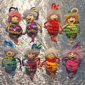 Op de afbeelding: Acht handgemaakte poppenornamenten met houten hoofden, garen lijfjes en kralenaccenten. Elke pop heeft een uniek kleurenschema, waaronder roze, blauw, groen en oranje. De poppen hebben geschilderde gezichten en pluizig haar.