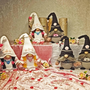 Könnte beinhalten: Sechs gehäkelte Zwergenfiguren mit spitzen Mützen, runden Nasen und Garnhaaren. Zwei Zwerge tragen weiße Mützen mit rosa Blumen, zwei tragen schwarze Mützen mit rosa Blumen und zwei haben herzförmige Akzente an Kleidung und Mützen.