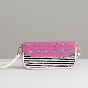 Puede incluir: Un bolso bandolera rectangular con una solapa rosa que presenta un patrón repetitivo de cebras plateadas. La parte inferior del bolso tiene rayas horizontales blancas y negras. Incluye una correa blanca para el hombro y herrajes dorados.