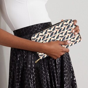 May include: A rectangular clutch bag featuring a cream-coloured background and a repeating pattern of black birds with red beaks. The bag has a gold zip and is held by a person wearing a white top and black skirt.