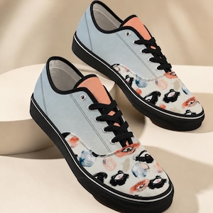 May include: A pair of low-top sneakers featuring a light blue upper, a peach-colored tongue, and black laces. The toe area displays a repeating pattern of stylized eyes and abstract watercolor-style shapes in black, red, and blue on a cream background.