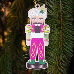 May include: Wooden Christmas ornament shaped like a nutcracker soldier. The ornament features a white turban, a black mustache, and a pink and green uniform. It hangs from a gold string against a backdrop of green pine needles.