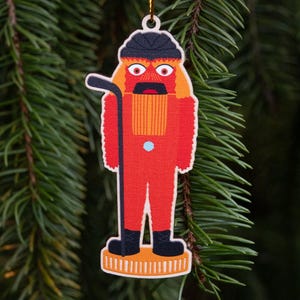May include: A red Christmas ornament shaped like a hockey player, with a black helmet and hockey stick. The ornament has orange hair and a black mustache. It is hanging from a gold string against a backdrop of green pine needles.
