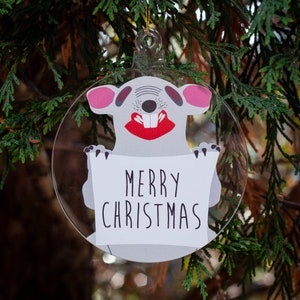 May include: Clear, round Christmas ornament featuring a cartoon mouse holding a sign that reads "MERRY CHRISTMAS". The mouse has pink ears, a red mouth, and a gray body. The ornament hangs from a gold string.