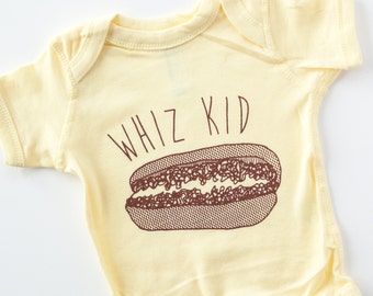 May include: A yellow baby onesie with a brown graphic of a hot dog and the text "WHIZ KID".