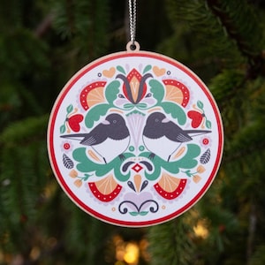 May include: Round Christmas ornament with a folk art design. The ornament features two black and white birds, surrounded by a floral pattern in red, green, and orange. The ornament is trimmed with a red border and has a silver string for hanging.