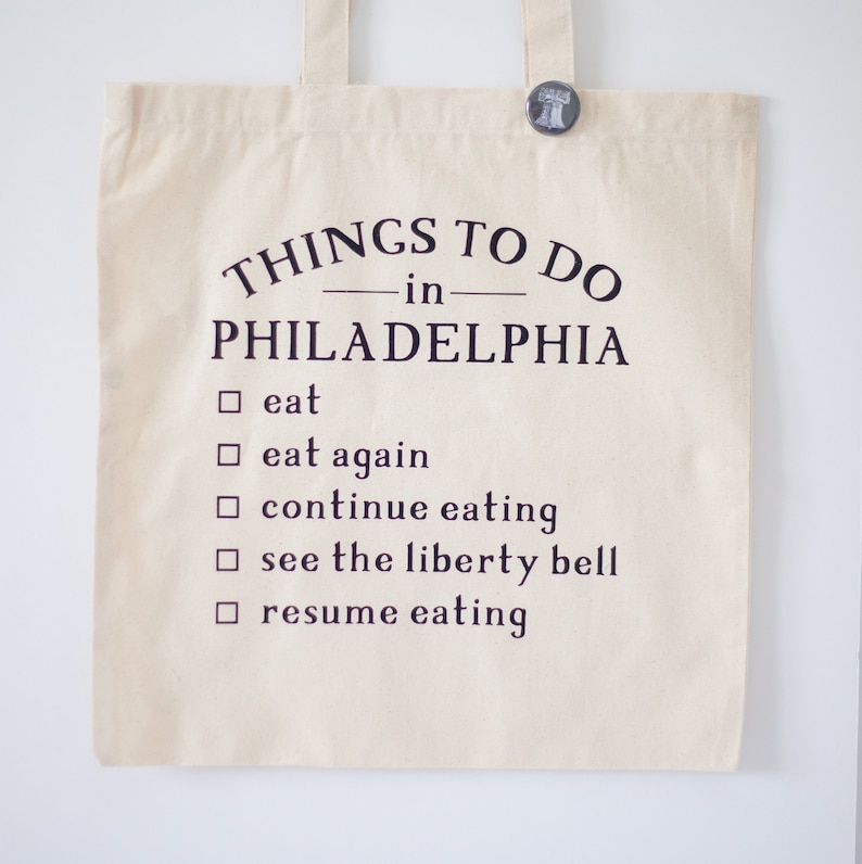May include: Cream-colored tote bag with the text "THINGS TO DO in PHILADELPHIA" and a checklist including "eat," "see the liberty bell," and "resume eating." A small button with a Liberty Bell design is pinned to the top.