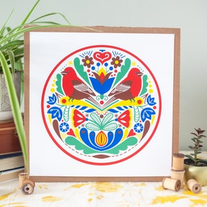 K&ouml;nnte beinhalten: Ein quadratischer Kunstdruck mit einem lebendigen, symmetrischen Design. Zwei rote V&ouml;gel blicken sich in einem kreisf&ouml;rmigen Rahmen an. Das Kunstwerk verwendet Rot, Blau, Gelb, Gr&uuml;n und Braun und schafft einen Volkskunststil. Der Druck ist vor einem wei&szlig;en Hintergrund.