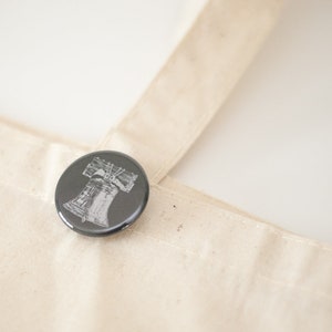 May include: A round, dark gray button with a detailed white illustration of the Liberty Bell. The button is pinned to a beige canvas tote bag. The image is well-lit, highlighting the button's design and the bag's texture.