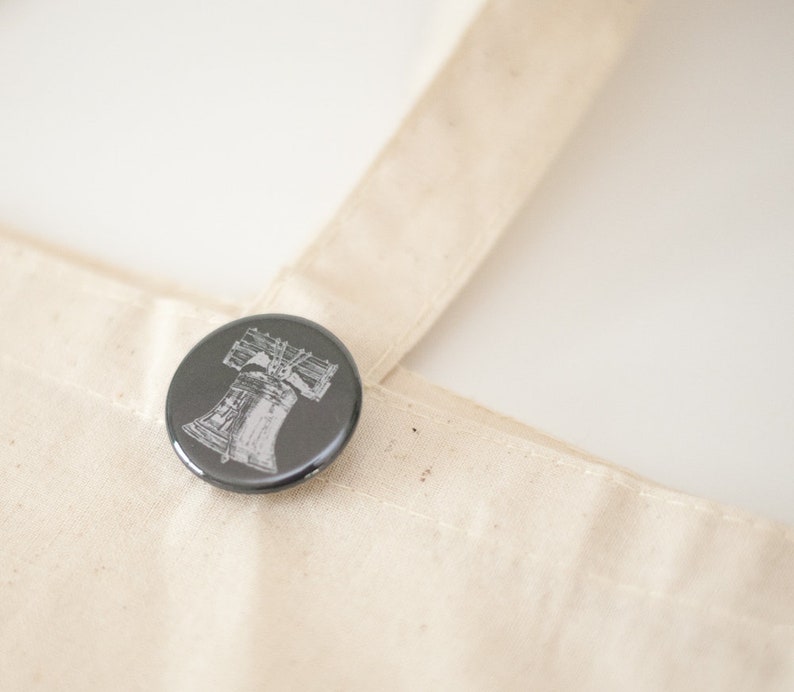May include: A round, dark gray button with a detailed white illustration of the Liberty Bell. The button is pinned to a beige canvas tote bag. The image is well-lit, highlighting the button's design and the bag's texture.