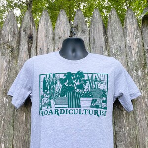 K&ouml;nnte beinhalten: Ein graues T-Shirt mit einer gr&uuml;nen Grafik einer Sammlung von Topfpflanzen. Der Text "HOARDICULTURIST" ist unter der Grafik gedruckt.