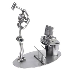 Op de afbeelding: Een metalen sculptuur van een persoon die met een hamer zwaait, naast een computerbureau met een monitor en toetsenbord staat. Het beeld is gemaakt van moeren, bouten en andere metalen onderdelen.