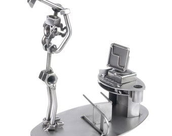 May include: A metal sculpture of a person swinging a hammer, standing next to a computer desk with a monitor and keyboard. The sculpture is made of nuts, bolts, and other metal pieces.