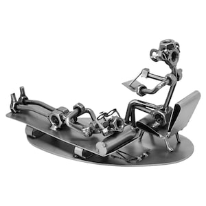 K&ouml;nnte beinhalten: Eine Metallskulptur aus zwei Figuren, eine sitzt und eine liegt, hergestellt aus Muttern, Schrauben und anderen Metallteilen. Die Figuren sind so positioniert, als w&auml;ren sie in einem Therapeutenb&uuml;ro, wobei die sitzende Figur einen Notizblock h&auml;lt und die liegende Figur eine Brille tr&auml;gt.