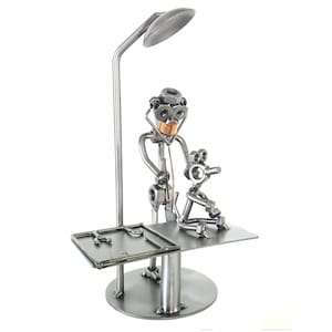 Puede incluir: Una escultura de metal de un médico examinando a un paciente. El médico lleva un estetoscopio y una lámpara frontal. El paciente está sentado en una mesa de examen. La escultura está hecha de tuercas, tornillos y otras piezas de metal.