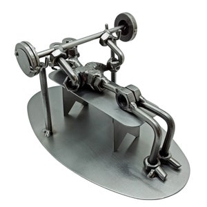 May include: A metal sculpture of a person lifting weights on a bench. The sculpture is made of nuts, bolts, and other metal pieces. The sculpture is on a metal base.