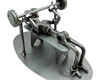 May include: A metal sculpture of a person lifting weights on a bench. The sculpture is made of nuts, bolts, and other metal pieces. The sculpture is on a metal base.