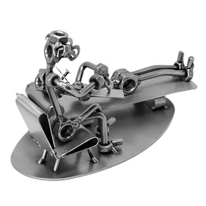 K&ouml;nnte beinhalten: Eine Metallsculptur, die einen Arzt zeigt, der einen Patienten untersucht. Der Arzt und der Patient sind aus Muttern, Schrauben und anderen Metallteilen gefertigt. Die Skulptur steht auf einem Metallsockel.