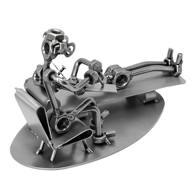 K&ouml;nnte beinhalten: Eine Metallsculptur, die einen Arzt zeigt, der einen Patienten untersucht. Der Arzt und der Patient sind aus Muttern, Schrauben und anderen Metallteilen gefertigt. Die Skulptur steht auf einem Metallsockel.