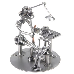 Op de afbeelding: Een metalen sculptuur van een dokter die een patiënt onderzoekt. De dokter is gemaakt van moeren, bouten en andere metalen onderdelen. De patiënt is ook van metaal en zit op een stoel. De dokter houdt een gereedschap in zijn hand en kijkt naar het hoofd van de patiënt. De sculptuur staat op een ronde metalen basis.