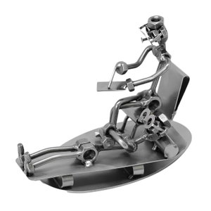 K&ouml;nnte beinhalten: Eine Metallsculptur einer Person, die auf einem Stuhl sitzt und die Beine ausgestreckt hat. Die Skulptur besteht aus Muttern, Schrauben und anderen Metallteilen. Die Person tr&auml;gt eine Brille und h&auml;lt ein Werkzeug in der Hand.