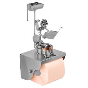 May include: A metal toilet paper holder featuring a figurine of a person sitting on a toilet and reading a book. The holder is made of silver metal and has a copper accent. The toilet paper roll is white.