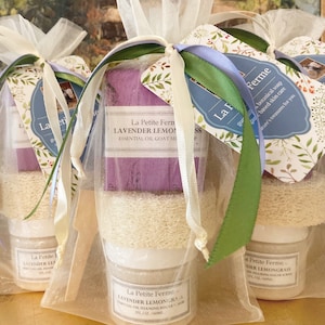 May include: Three gift bags filled with lavender and lemongrass scented bath products. Each bag contains a bar of soap, a jar of foaming sugar scrub, and a loofah. The bags are tied with white ribbon and decorated with floral patterned tags that read "La Petite Ferme".