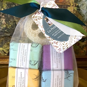May include: A gift set of goat milk soaps in a sheer bag, tied with green and teal ribbons. The set includes bars in various colors and scents, such as eucalyptus peppermint and almond & Florida honey. A natural loofah sponge and a tag with the brand name are included.