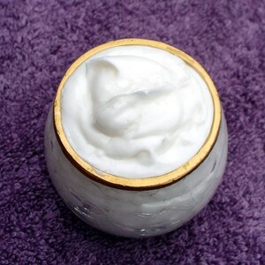 May include: A white cream in a glass jar with a gold rim. The cream is thick and fluffy, and the jar is sitting on a purple towel.