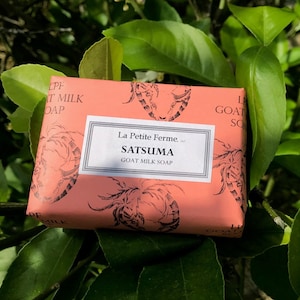 May include: A bar of Satsuma goat milk soap wrapped in peach-colored paper. The paper is printed with a repeating pattern of a goat's head and the words "La Petite Ferme" and "GOAT MILK SOAP". The soap is surrounded by green leaves.