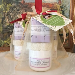 May include: A gift set with a white jar of foaming sugar body polish and a bar of goat milk soap, both labeled "La Petite Ferme Blackberries & Vanilla". The set is packaged in a sheer, gold organza bag with a red ribbon.