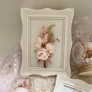 Könnte beinhalten: Ein gerahmter Trockenblumen-Arrangement mit rosa und weißen Blumen und getrockneten Gräsern. Der Rahmen ist weiß mit einem dekorativen Rand.