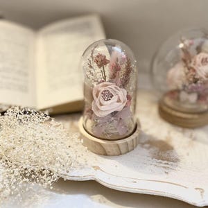 Peut inclure: Un dôme en verre avec une base en bois contenant des fleurs séchées roses et blanches. Le dôme est posé sur une surface en bois blanc.