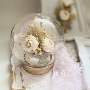 Könnte beinhalten: Eine Glaskuppel mit einem Holzsockel, die getrocknete weiße Blumen und Grün enthält. Die Kuppel steht auf einer weißen Oberfläche mit einer Spitzenüberlage.