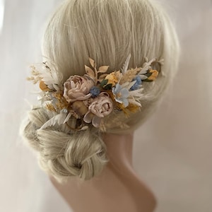 Könnte beinhalten: Ein Blumenhaarkamm mit getrockneten Blumen in Beige-, Braun-, Blau- und Goldtönen. Der Kamm ist aus getrockneten Blumen und Grünzeug gefertigt, darunter Federn und Blätter.