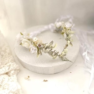 Puede incluir: Una corona floral blanca con delicadas flores blancas y hojas verdes. La corona está sobre una superficie blanca.