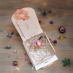 Könnte beinhalten: Eine weiße Geschenkbox mit einem goldenen Armband und einem Blumenarrangement. Die Box enthält ein Etikett mit dem Text "Will You Be My Bridesmaid?" und ein rosa Band. Auf einer Holzoberfläche sind Blumen in Lila, Rosa und Gelb verstreut.