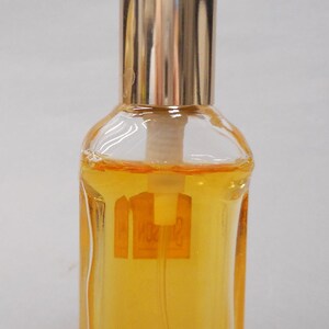 Op de afbeelding: Een kleine, heldere glazen parfumfles met een goudkleurige dop en een gouden vloeistof erin. De fles heeft een rechthoekige vorm met een licht afgeronde bovenkant en een goudkleurige dop. De fles heeft een label met tekst.