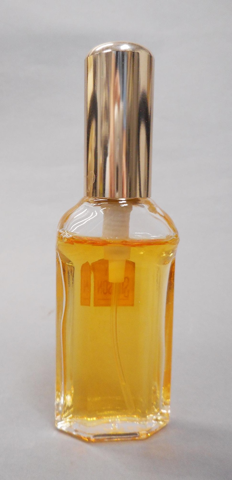 Op de afbeelding: Een kleine, heldere glazen parfumfles met een goudkleurige dop en een gouden vloeistof erin. De fles heeft een rechthoekige vorm met een licht afgeronde bovenkant en een goudkleurige dop. De fles heeft een label met tekst.