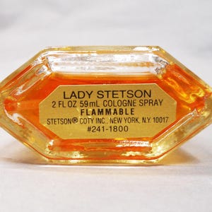 Puede incluir: Un frasco de perfume transparente, octogonal, con l&iacute;quido dorado en su interior. La etiqueta dice "LADY STETSON 59ml COLOGNE SPRAY FLAMMABLE". El frasco es de STETSON COTY INC., Nueva York, N.Y. #241-1800.