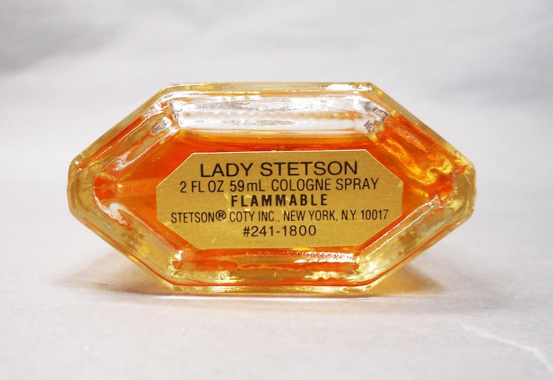 Puede incluir: Un frasco de perfume transparente, octogonal, con l&iacute;quido dorado en su interior. La etiqueta dice "LADY STETSON 59ml COLOGNE SPRAY FLAMMABLE". El frasco es de STETSON COTY INC., Nueva York, N.Y. #241-1800.