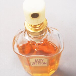 Puede incluir: Un frasco de perfume Lady Stetson vintage. El frasco de vidrio transparente tiene forma octogonal y contiene un l&iacute;quido naranja dorado. El frasco tiene un cuello dorado y una tapa color crema. La etiqueta dice "Lady Stetson".